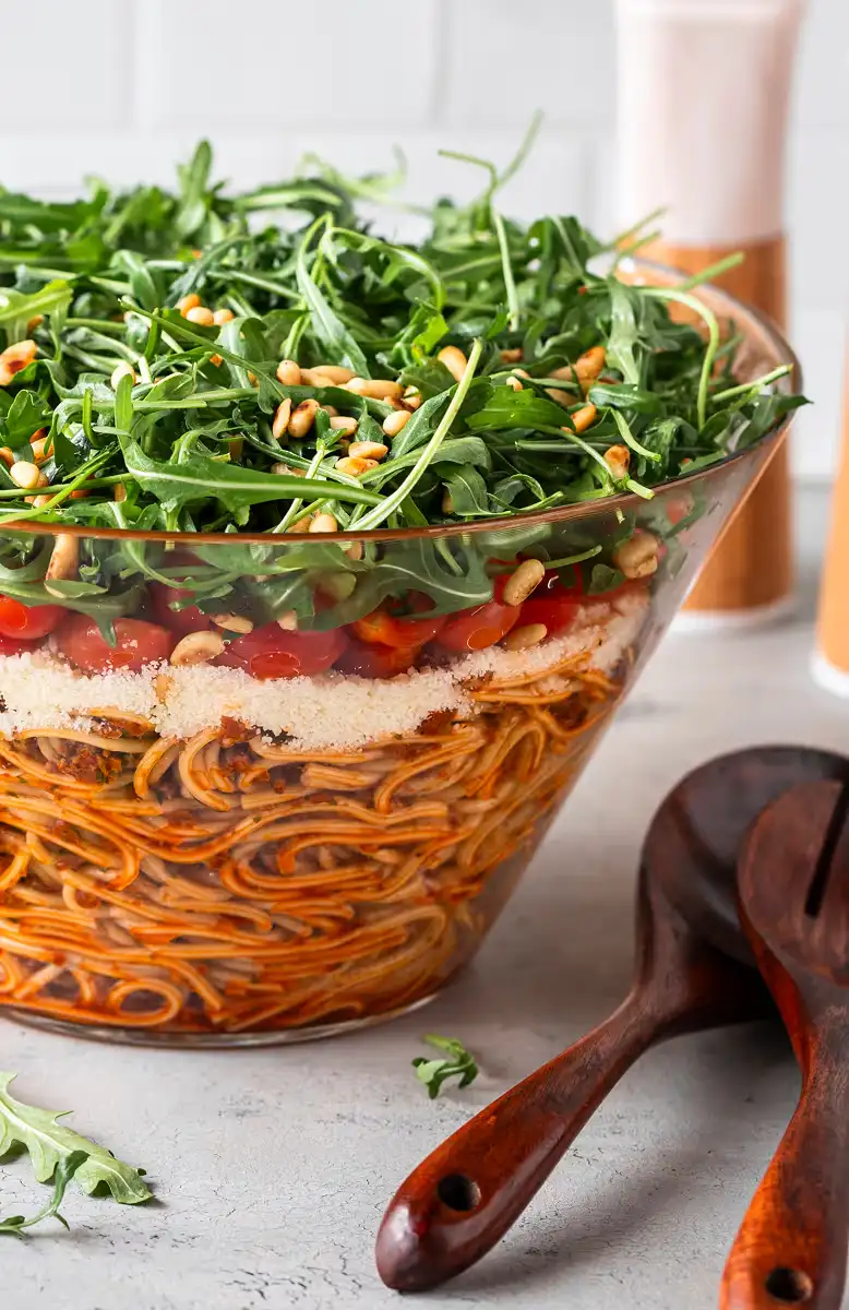 Layered spaghetti salad in a glass bowl topped with arugula and pine nuts, with wooden salad utensils beside it.