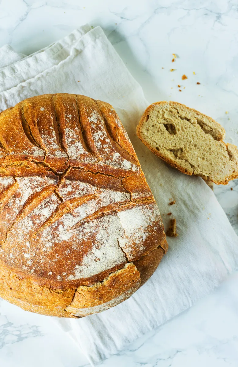 Freshly baked round loaf of bread with a slice cut, resting on a wooden cutting board with crumbs scattered around.