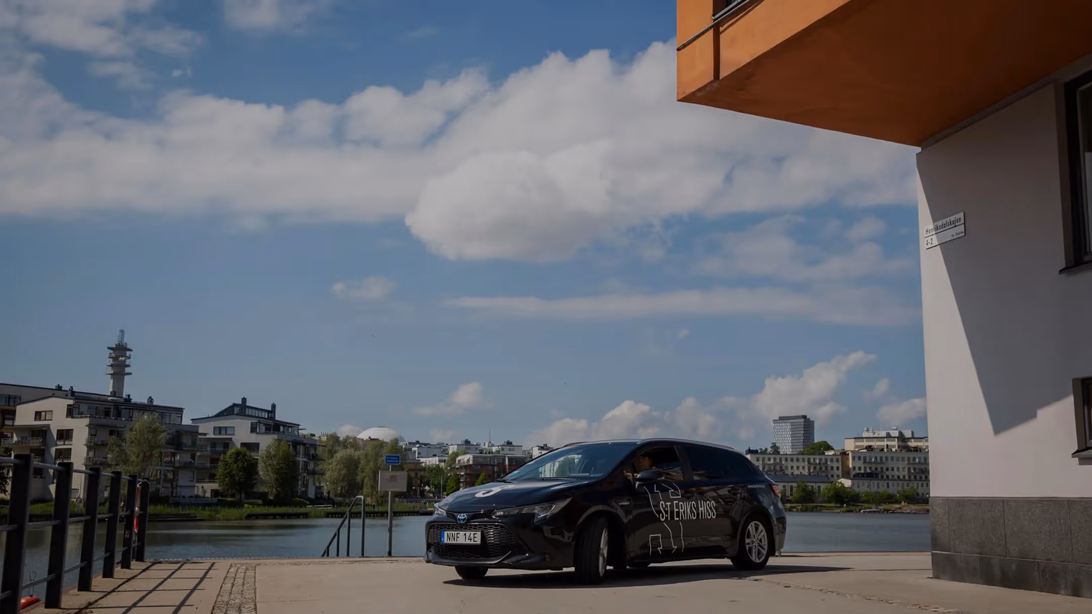Svart servicebil från S:t Eriks Hiss parkerad vid vattnet med stadens byggnader i bakgrunden.