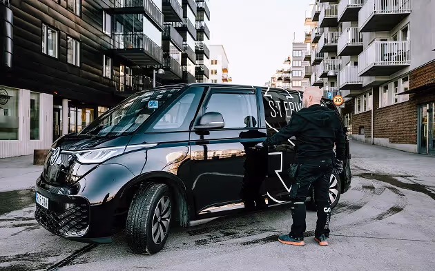 Man står bredvid en svart servicebil med text, med stadsbebyggelse i bakgrunden.