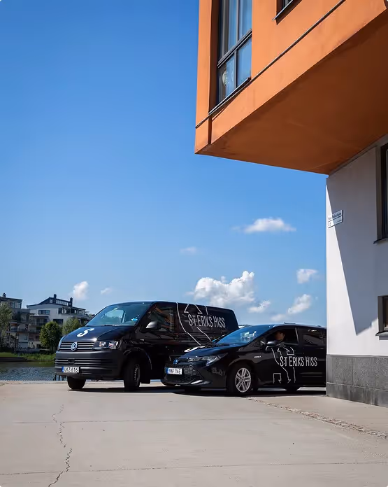 Två svarta servicebilar från S:t Eriks Hiss parkerade vid en orange och vit byggnad en solig dag.