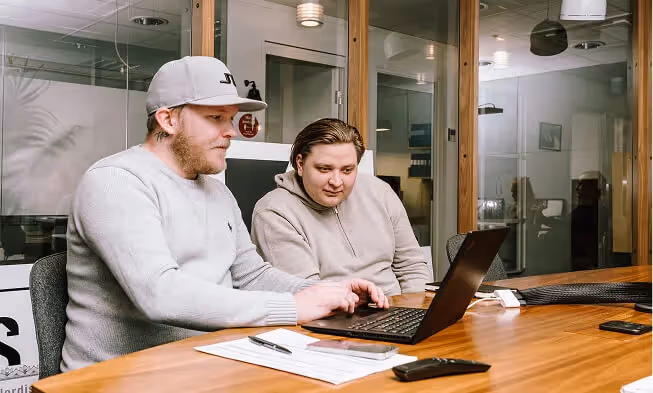 Två män sitter vid ett träbord och arbetar vid en laptop i ett modernt kontor med glasväggar.