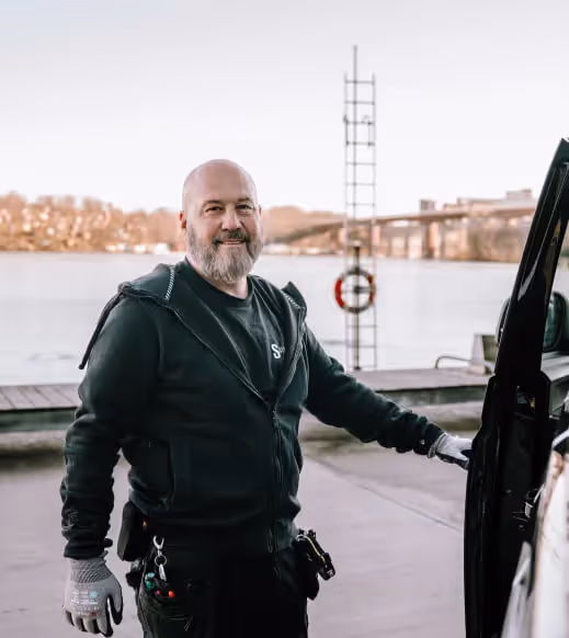 Leende tekniker i svart jacka och handskar håller i en bildörr vid vattnet med en bro i bakgrunden.