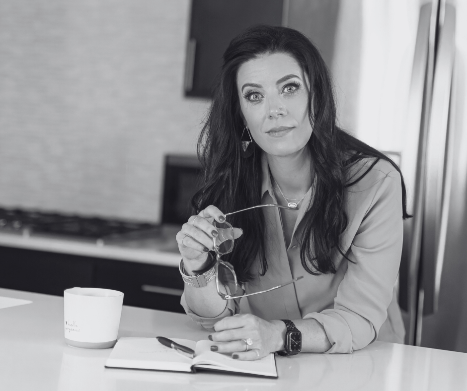 Amy Reczek seated at a kitchen counter with a notebook and coffee mug, holding reading glasses