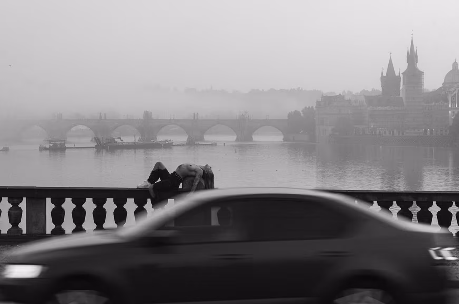 Černobílá fotografie muže bez trička, který leží na zábradlí mostu nad řekou za projíždějícím autem, s mlhavým městským panoramatem v pozadí.