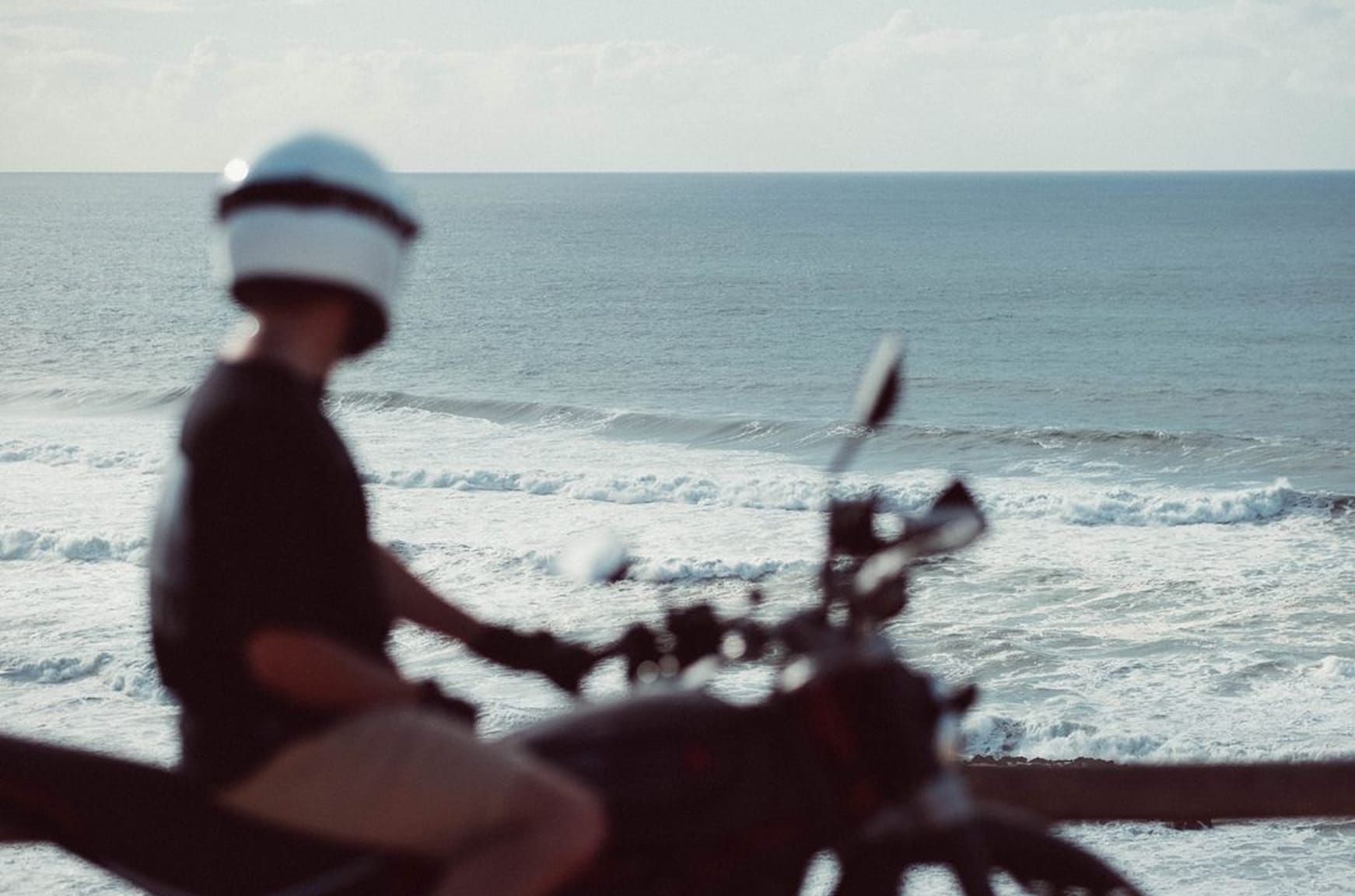 Zaměřená fotografie osoby s bílou helmou na motocyklu u moře s vlnami v pozadí.
