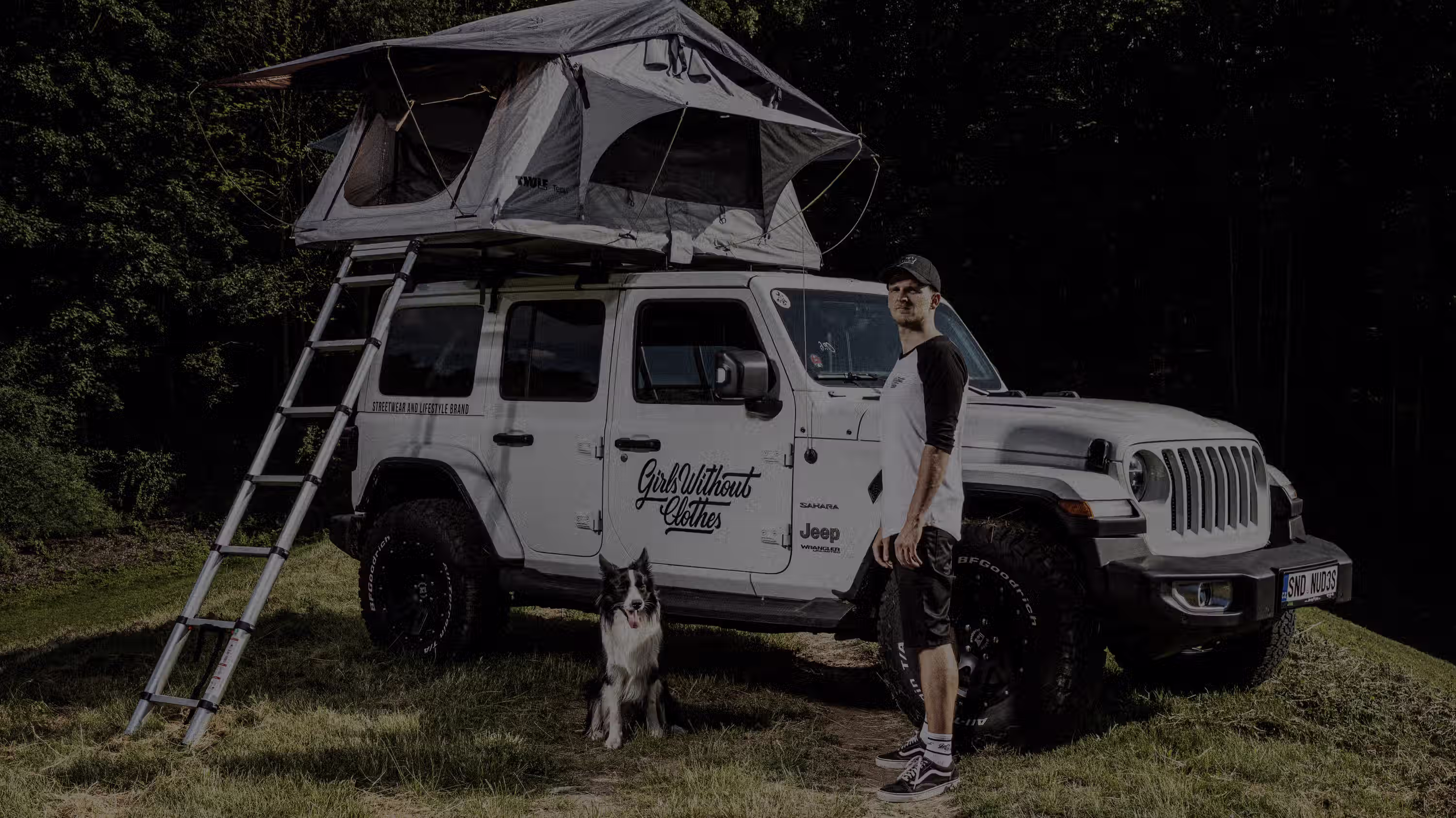 Muž stojící vedle bílé Jeep Wrangler s rozloženým střešním stanem a černobílý pes sedící na trávě.