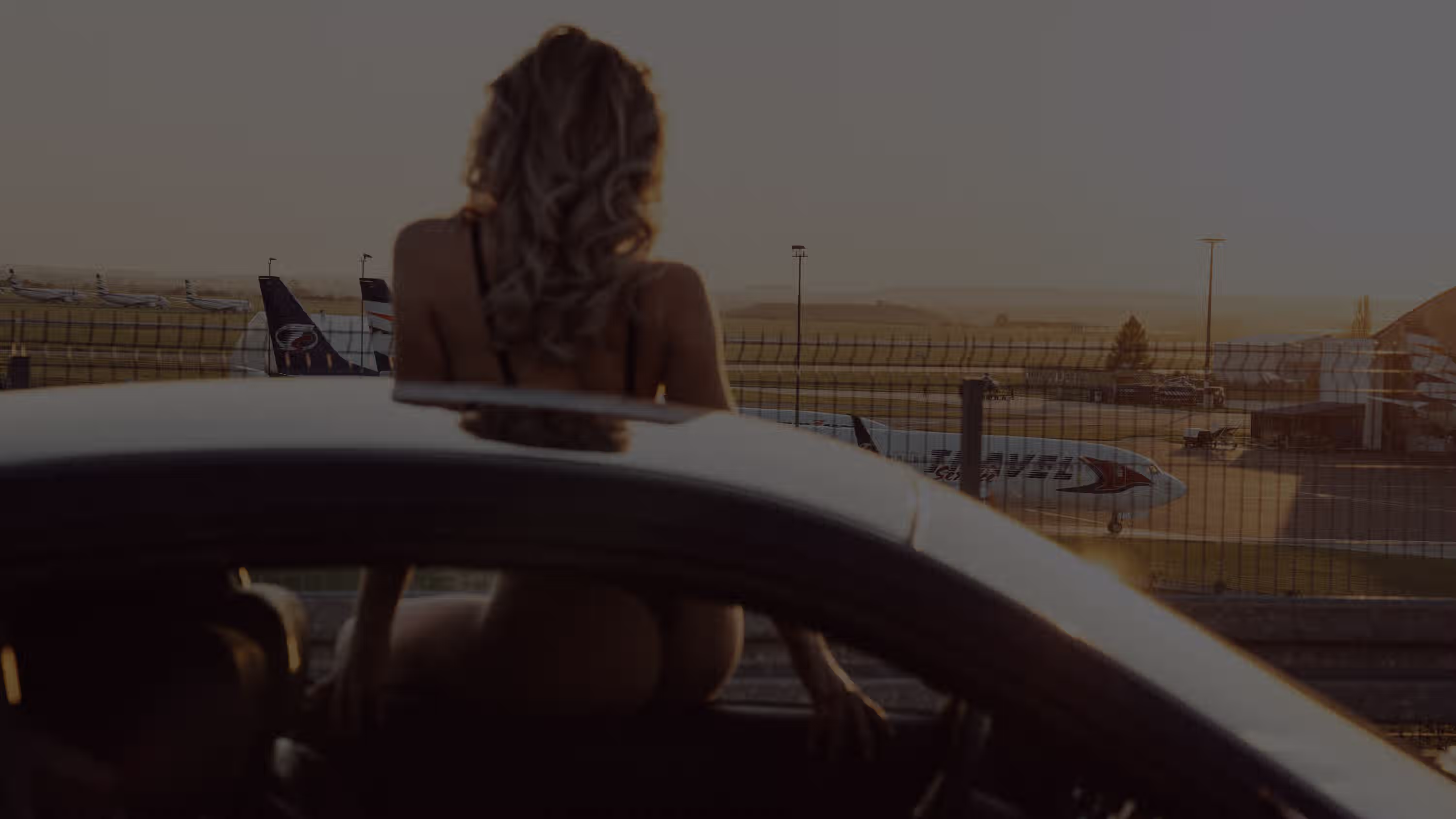 Žena s vlnitými vlasy sedí v autě u okna a dívá se na letadla na letišti za plotem za soumraku.