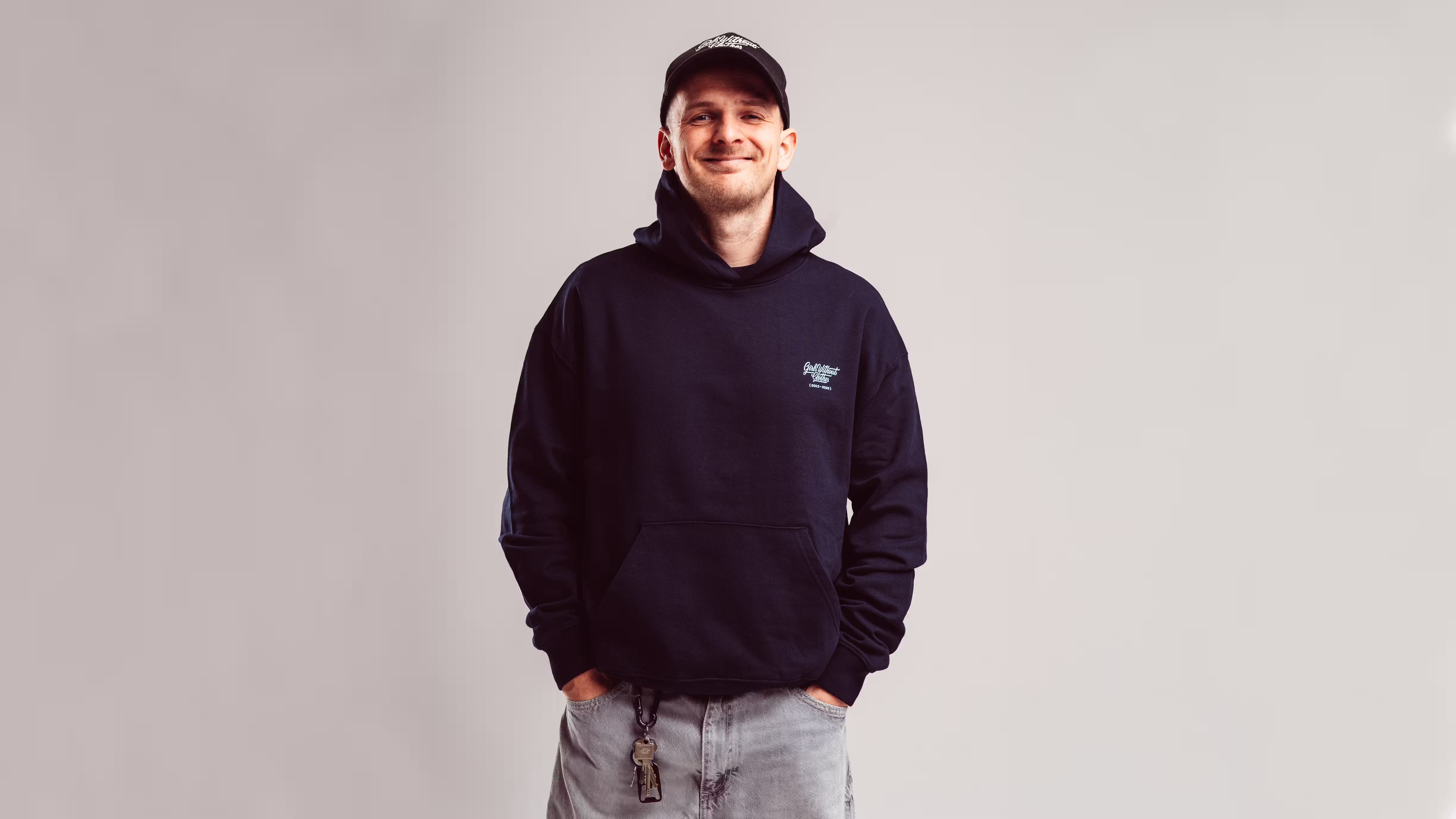 Muž v černé mikině s kapucí a šedých džínách stojí s rukama v kapsách a usmívá se.