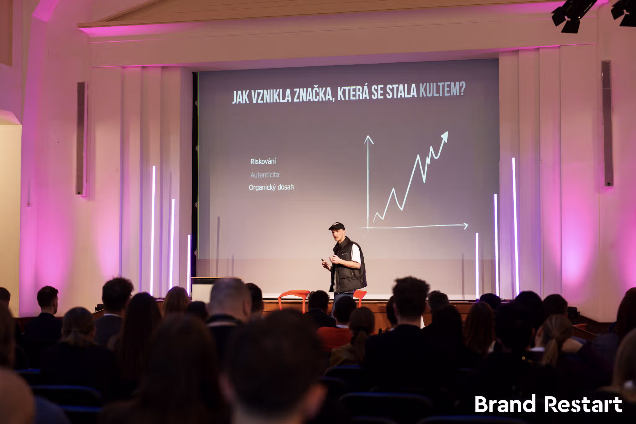 Muži na pódiu před projekcí grafu s názvem ‚Jak vznikla značka, která se stala kultem?‘ s tématy riskování, autenticity a organického dosahu, před publikem v místnosti s růžovým osvětlením.