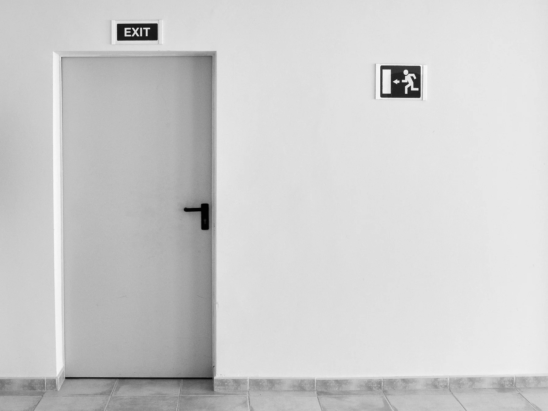 Closed gray exit door on white wall with black exit sign above and running exit icon to the right.