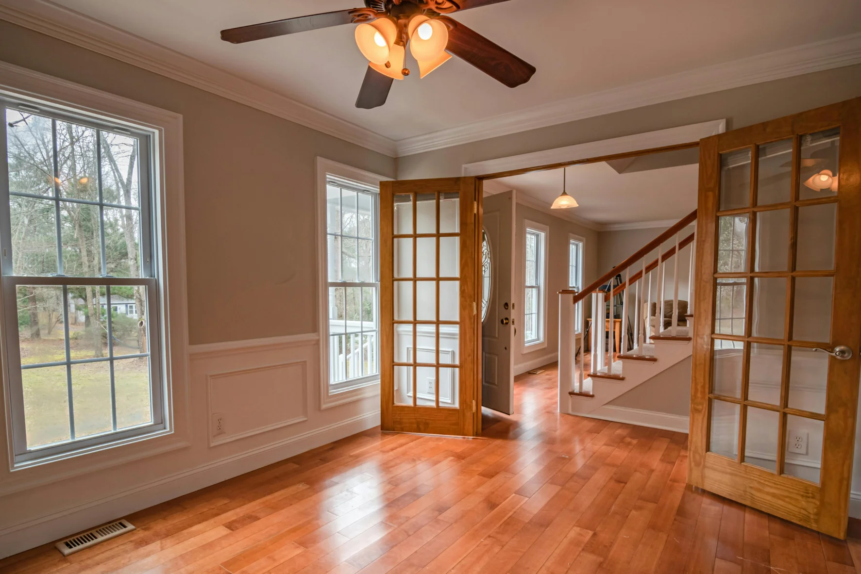 Empty room with wooden floor, two large windows, ceiling fan with lights, and open wooden French doors leading to a hallway with stairs.