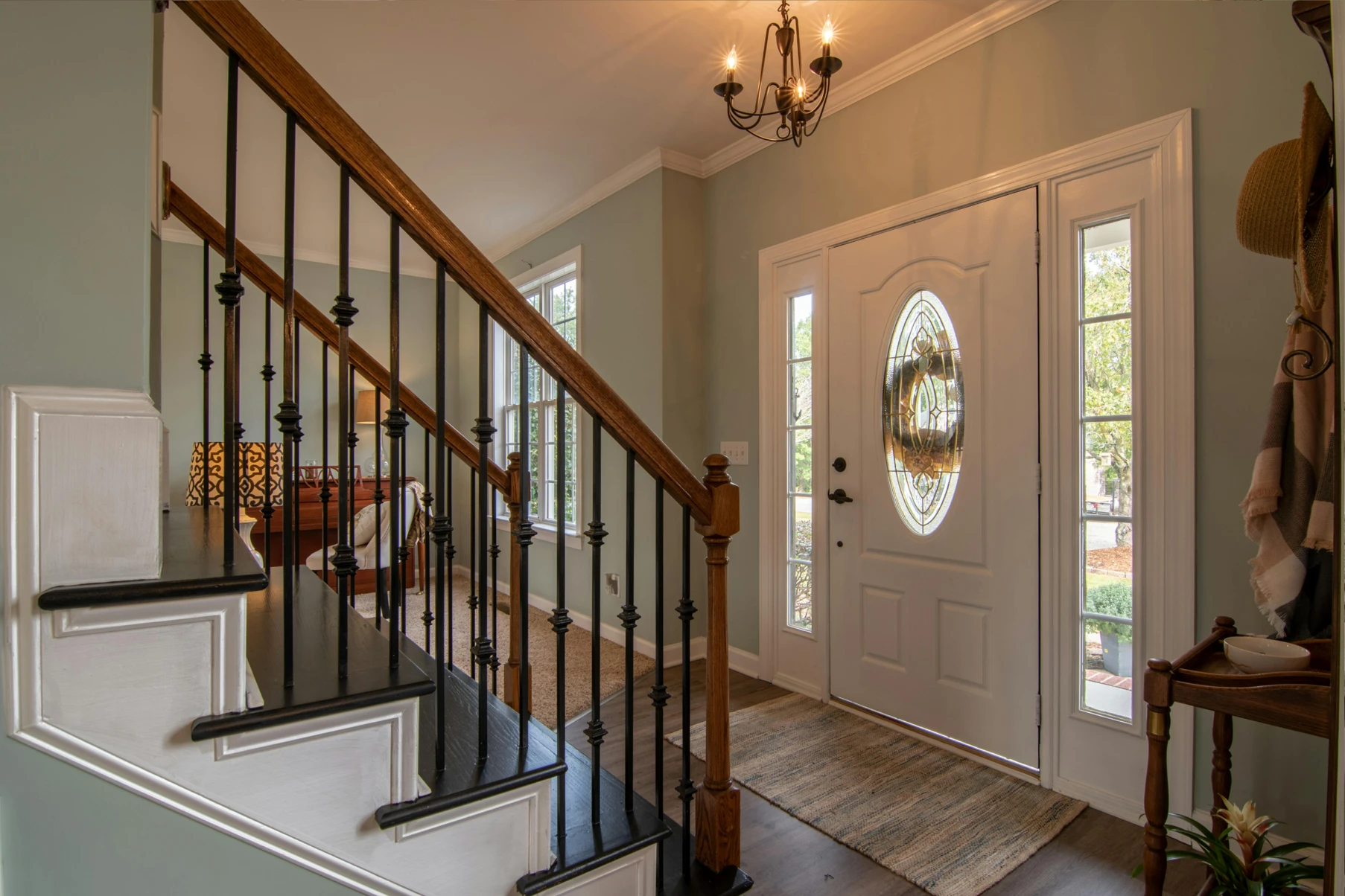 Interior view of a home entryway featuring a white front door with decorative oval glass and sidelights, a wooden staircase with black iron balusters, a chandelier, and a small wooden table with hats and a scarf.