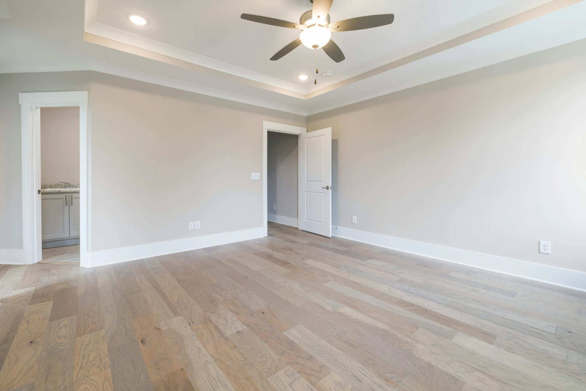 Empty room with light gray walls, light wood flooring, white trim, a ceiling fan with light, an open door, and entrance to a bathroom with a granite countertop.