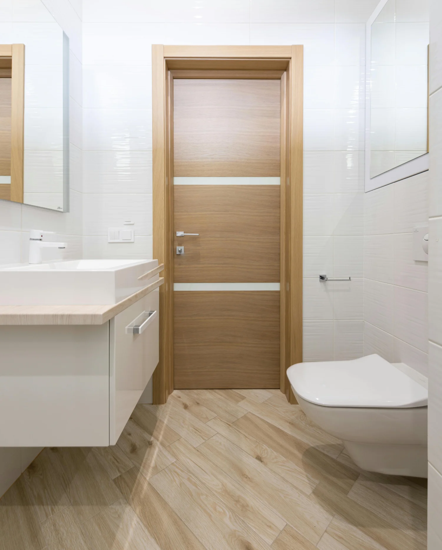 Modern bathroom interior with a light oak door, white sink vanity, and wall-mounted toilet on wood-patterned tile floor.