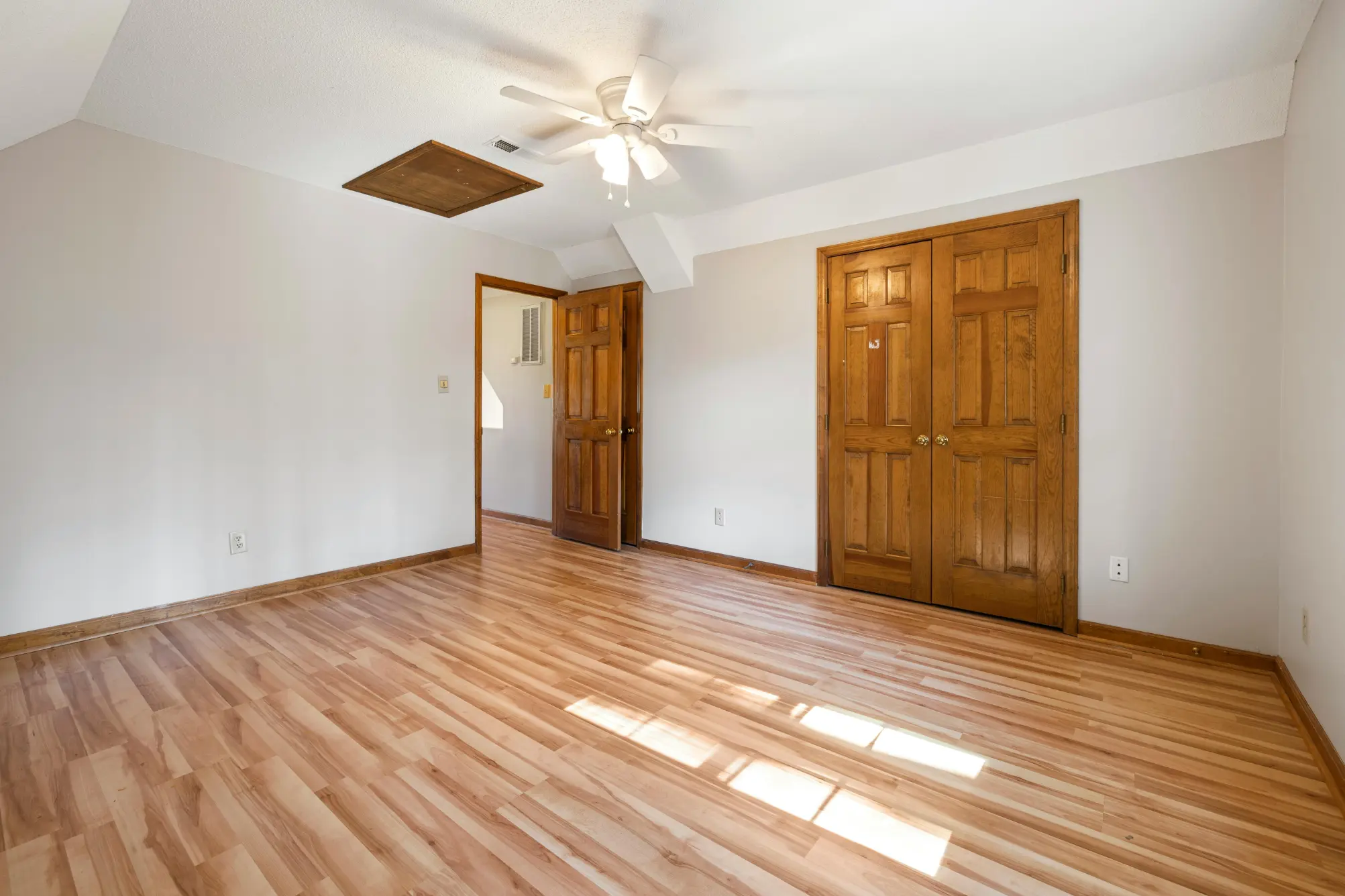 Empty room with light wood flooring, white walls, a wooden door slightly open, a closed wooden double closet door, and a ceiling fan with light.