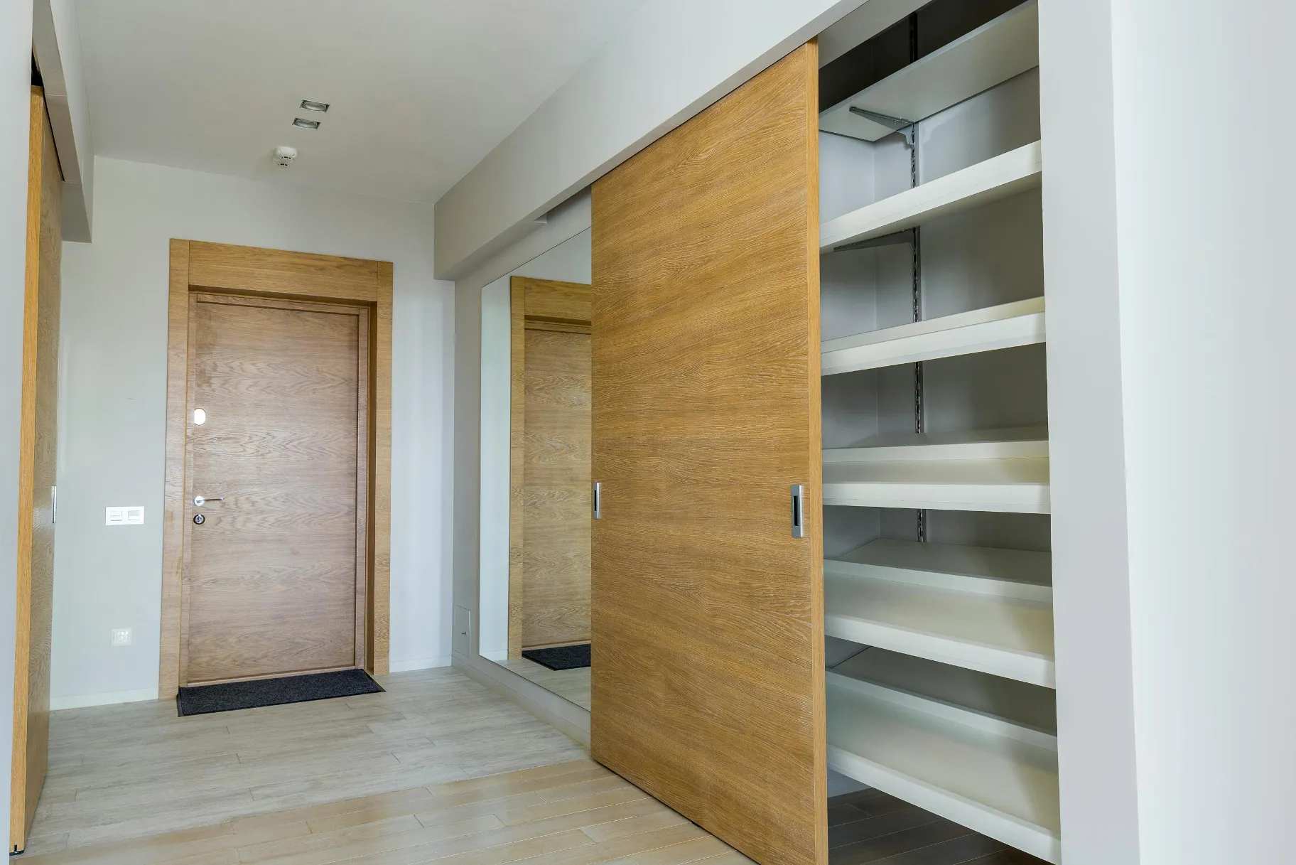 Interior hallway with wooden sliding door partially open revealing empty white shelves and a closed wooden entry door with a black doormat.