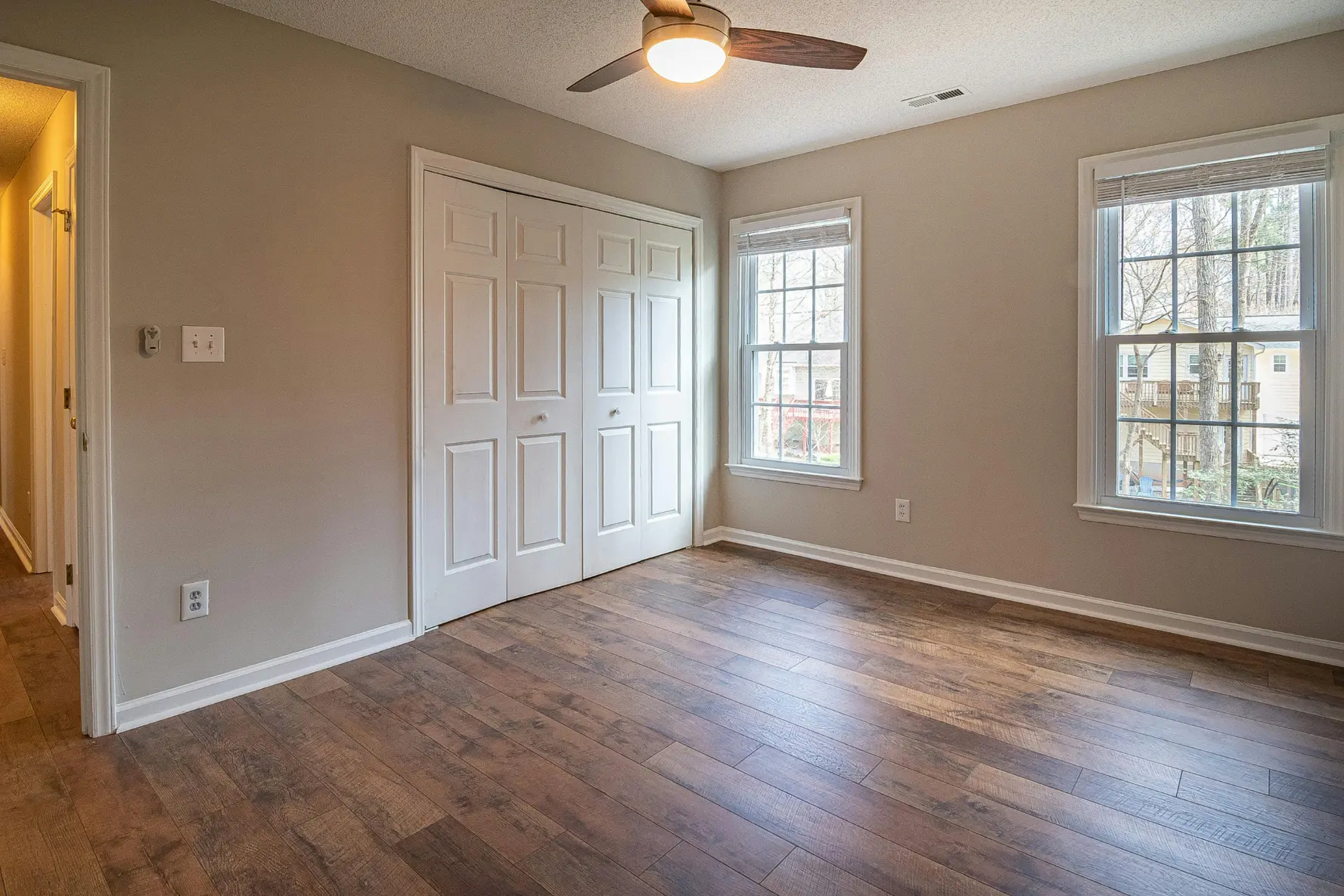 Empty room with wooden floor, two large windows, white double closet doors, and ceiling fan with light.