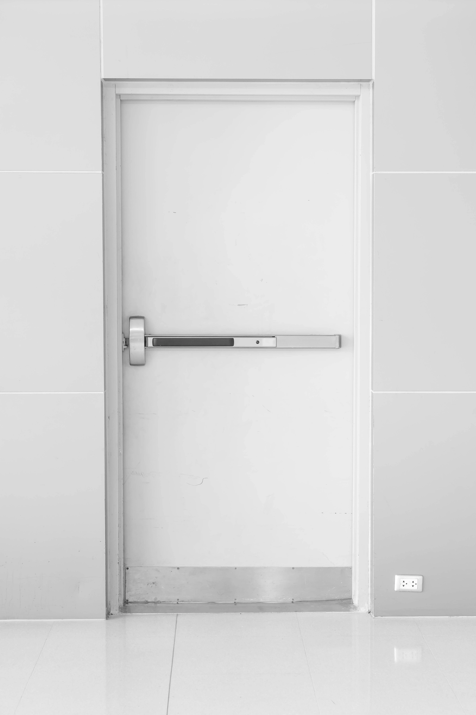 White fire exit door with a horizontal push bar and a metal plate at the bottom.