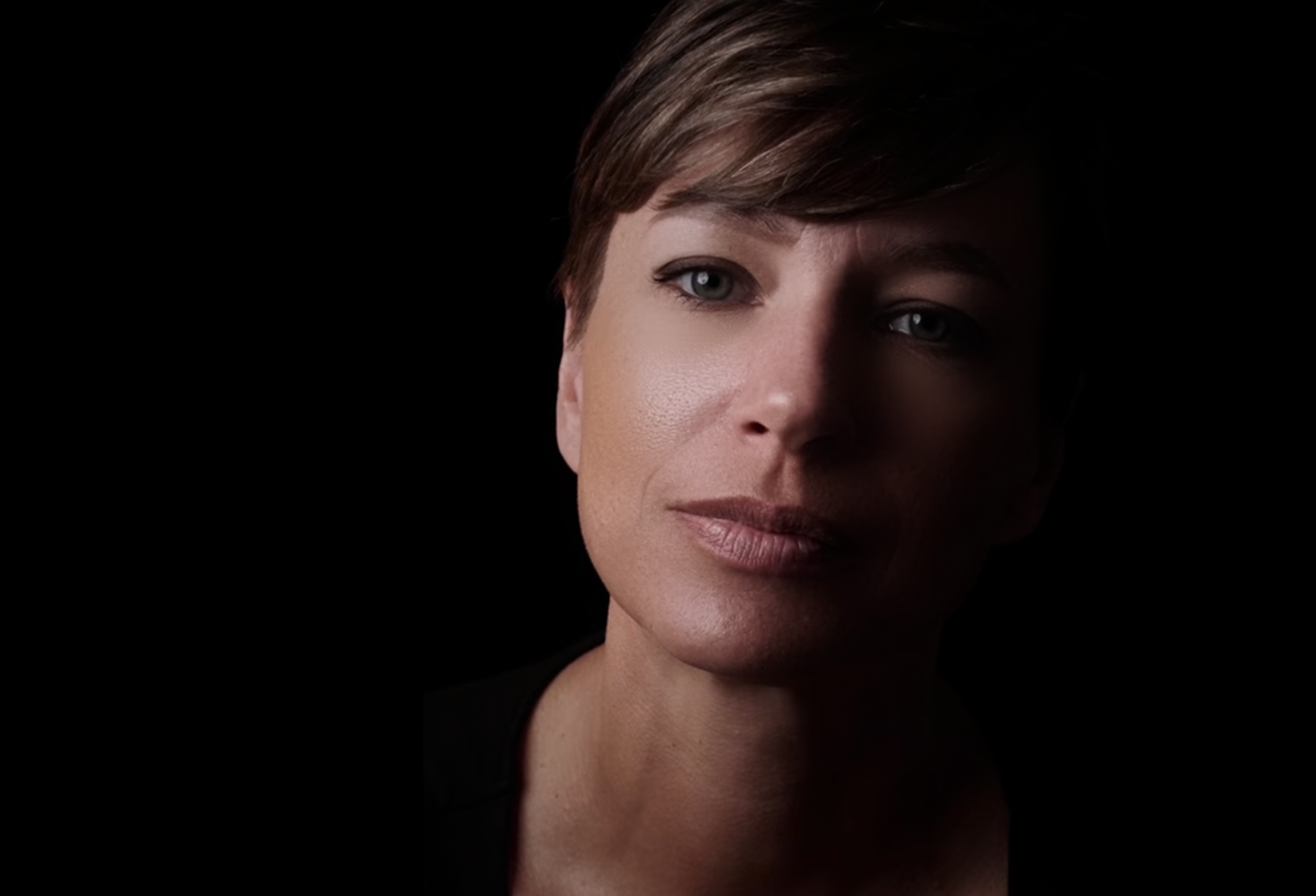 Close-up portrait of a woman with short brown hair and light makeup against a black background.