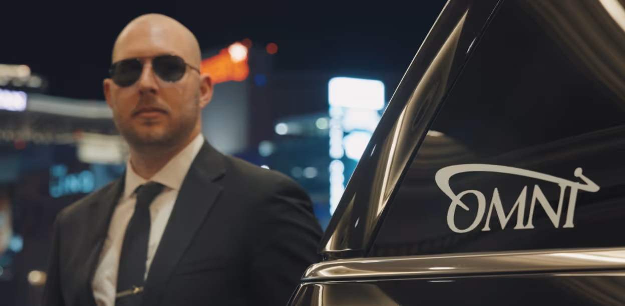 A private Omni chaffeur in a black suit and tie wearing sunglasses standing next to a black car with an OMNI logo on the window at night.