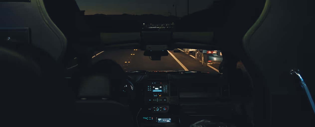 View from inside a car looking out at a dark road at night with parked cars on the right side.