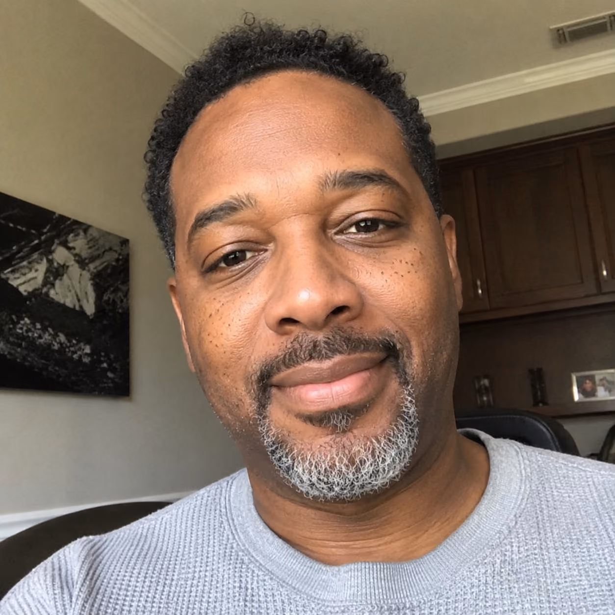 A man with short curly hair and a salt-and-pepper goatee wearing a gray sweater smiling slightly.