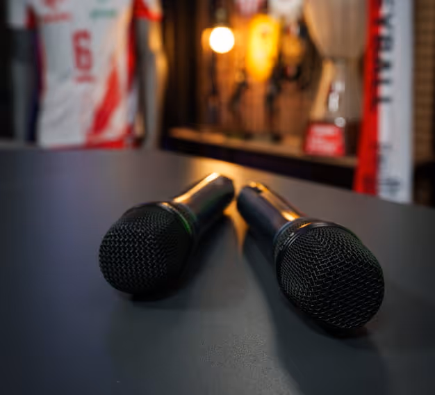 Dwa czarne mikrofony leżące na ciemnej powierzchni na tle rozmazanego stroju sportowego i trofeów.