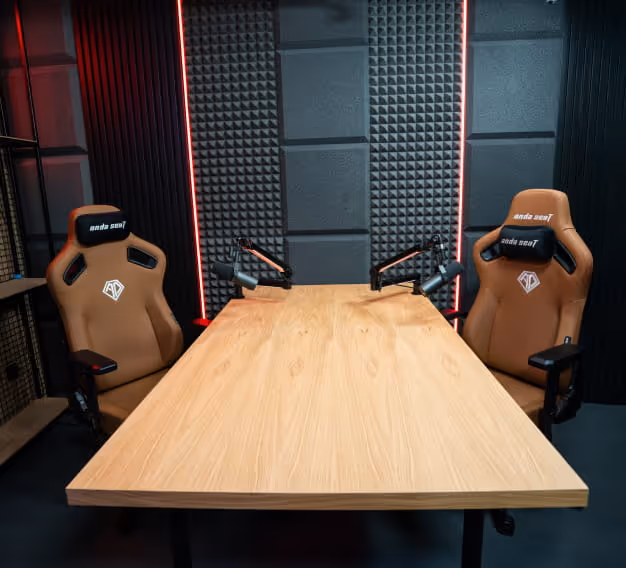 Dwa brązowe fotele w stylu gamingowym z czarnymi podłokietnikami mikrofonu po obu stronach jasnego drewnianego stołu w dźwiękoszczelnym pomieszczeniu nagraniowym z czerwonymi paskami oświetleniowymi LED.