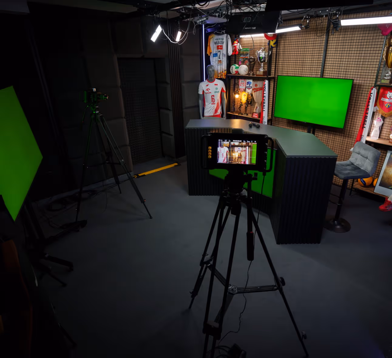 Studio wideo w pomieszczeniu z kamerami na statywach, manekinem w koszulce sportowej, zakrzywionym biurkiem, zielonymi ekranami i zamontowanym na ścianie telewizorem z zielonym ekranem.