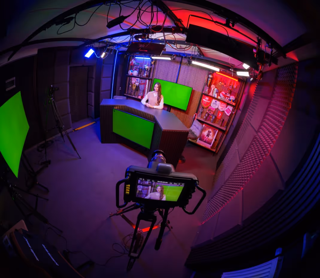 Szerokokątny widok studia nagrań z kobietą siedzącą za biurkiem z zielonymi ekranami i profesjonalnym sprzętem oświetleniowym.