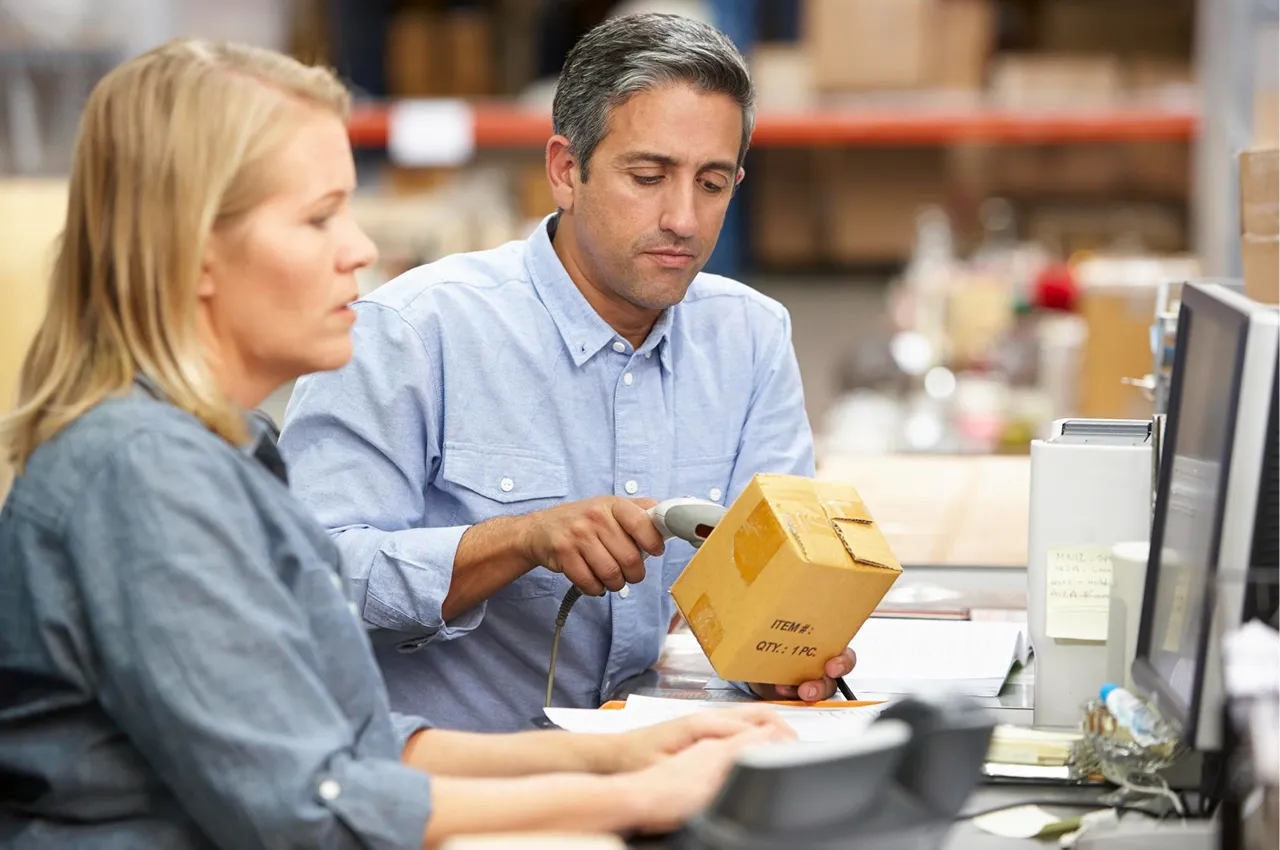 Man scanning a box with a barcode scanner while a woman works on a computer nearby in a warehouse office.