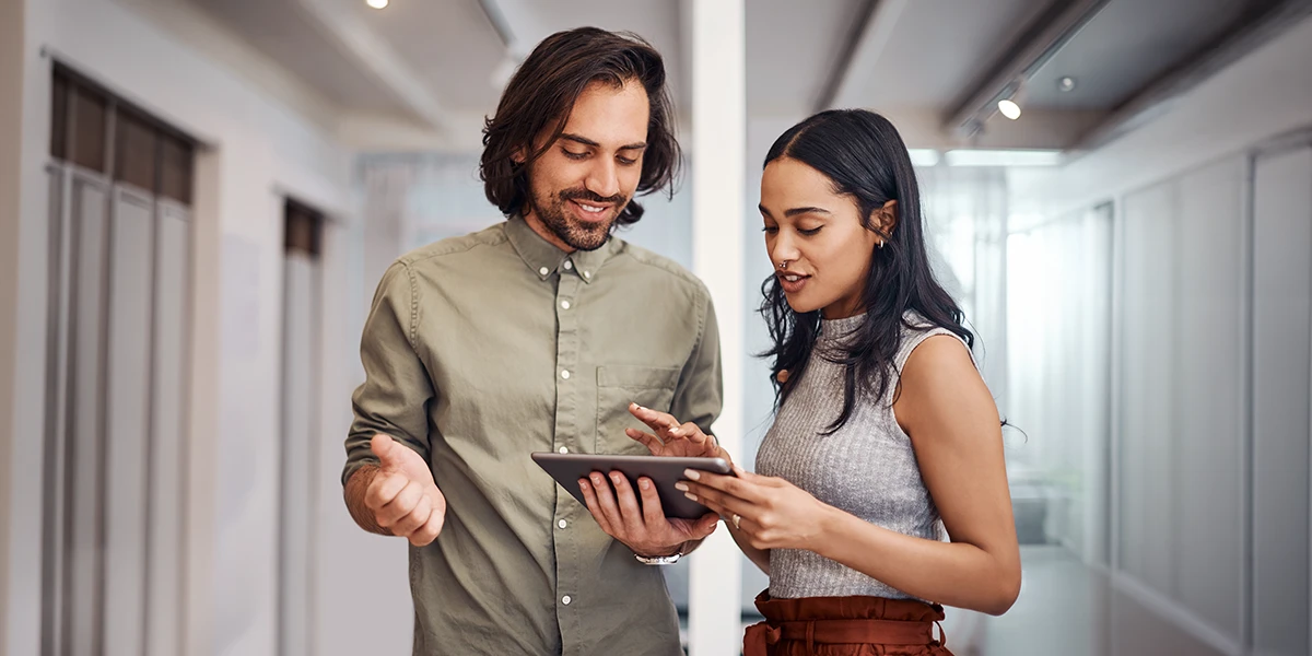 Two colleagues in a modern office hallway looking at and discussing content on a digital tablet, smiling and engaged.