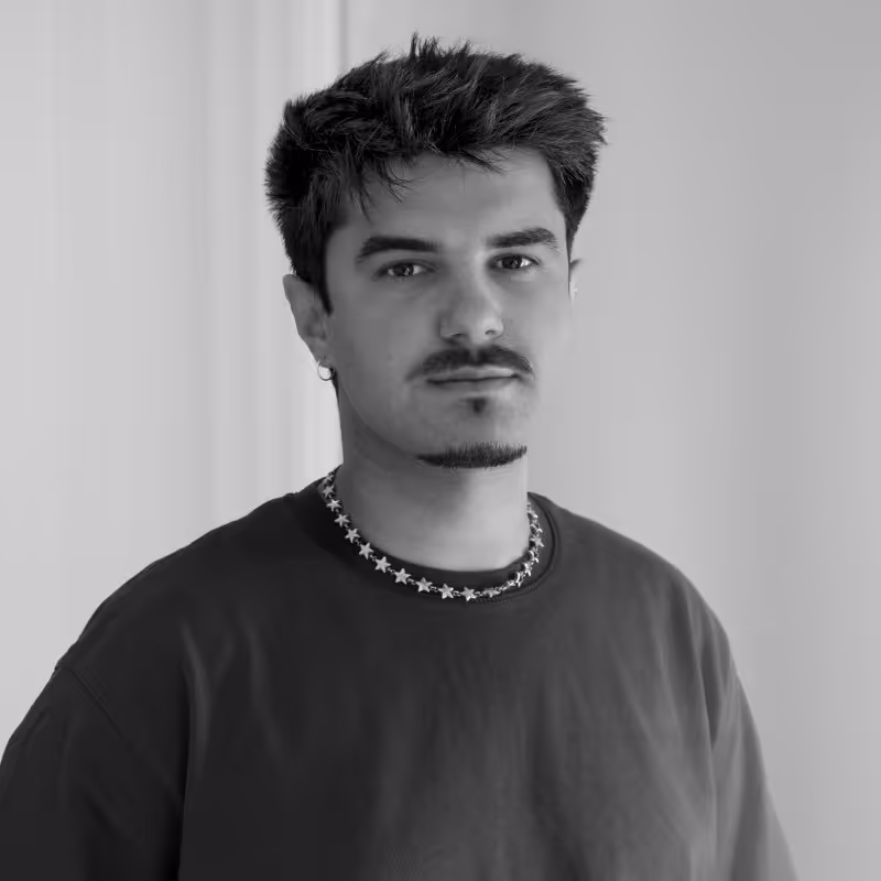 Retrato en blanco y negro de un hombre joven con peinado despeinado, barba corta, pendiente y collar de estrellas, que mira hacia la cámara.