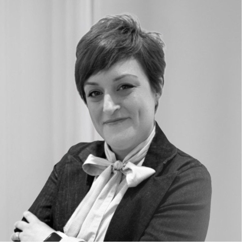 Mujer de cabello corto con camisa blanca y chaqueta oscura, sonriendo con los brazos cruzados.