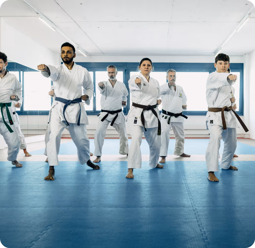 Group of six martial artists in white uniforms performing synchronized punches on blue mats in a bright training room.