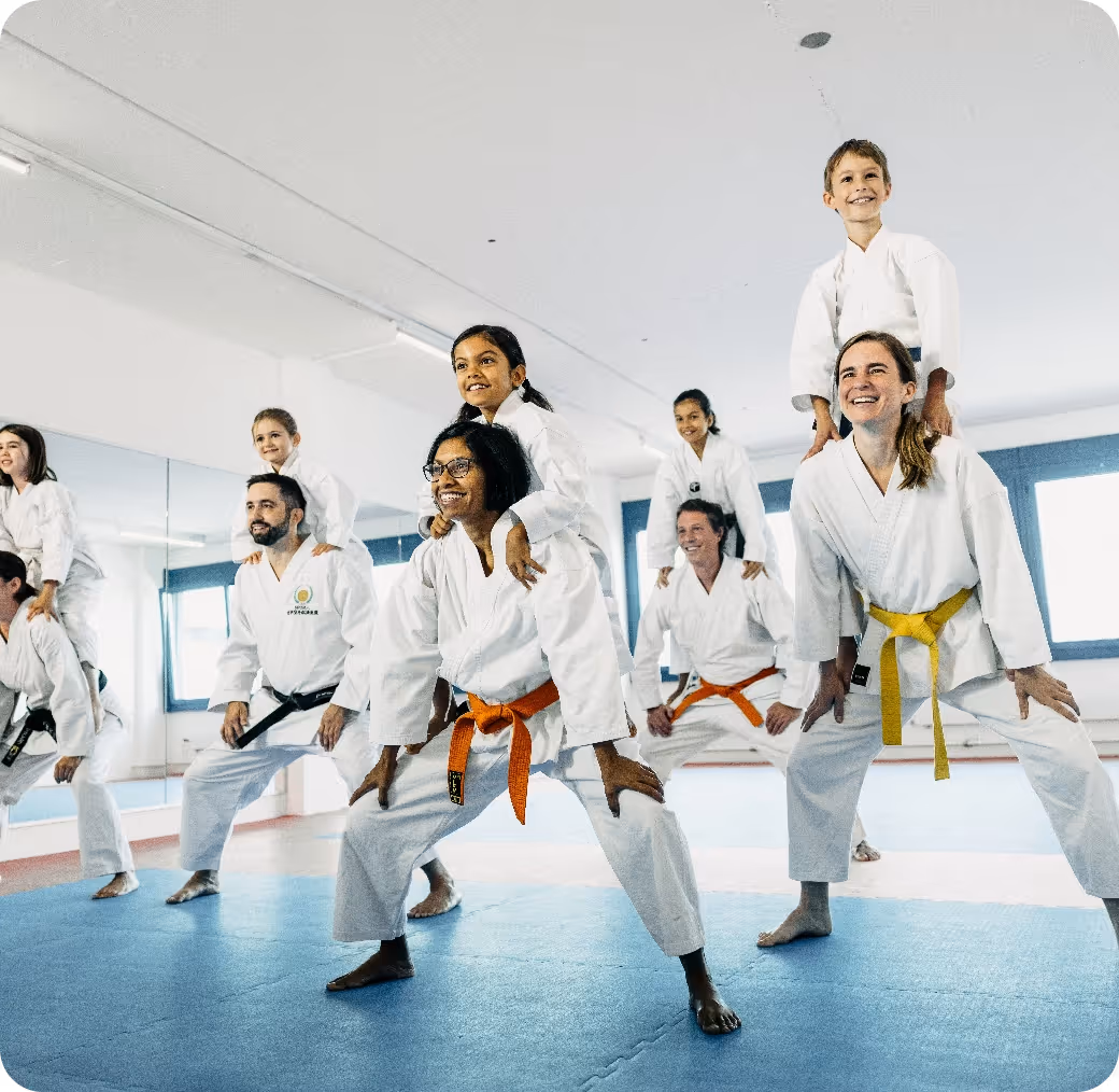 Adults in karate uniforms with colored belts carrying children on their backs inside a martial arts studio.