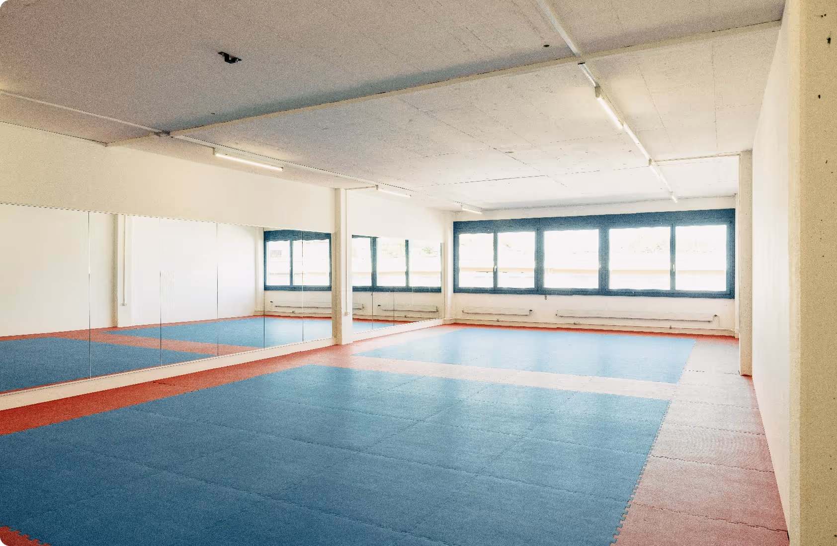 Empty martial arts training room with padded blue and red flooring and large mirrors on one wall.