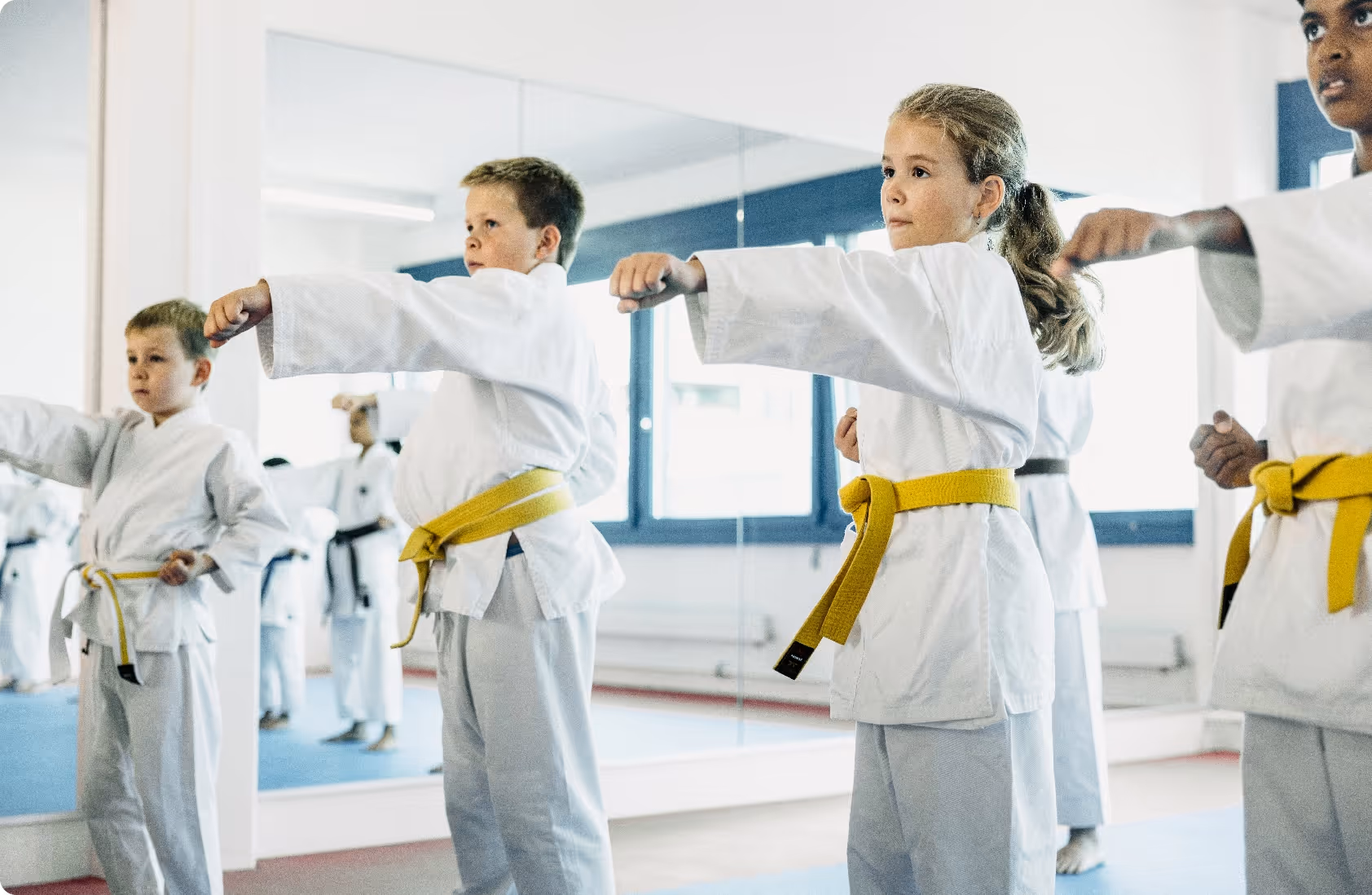 Children with yellow belts practicing martial arts punches in a dojo with mirrored walls.