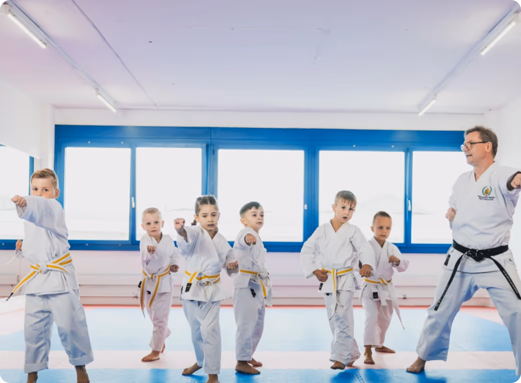 Martial arts instructor leads a group of young children with yellow belts practicing punch techniques in a bright dojo.