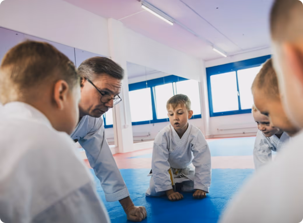 Martial arts instructor demonstrating a technique on the blue mat with young children in white uniforms gathered around.