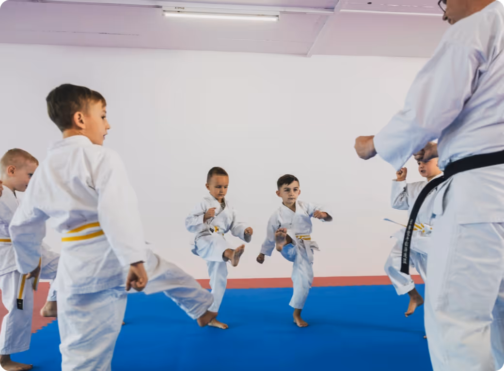 Young children in white karate uniforms with yellow belts practicing kicking on blue mats, guided by an instructor with a black belt.