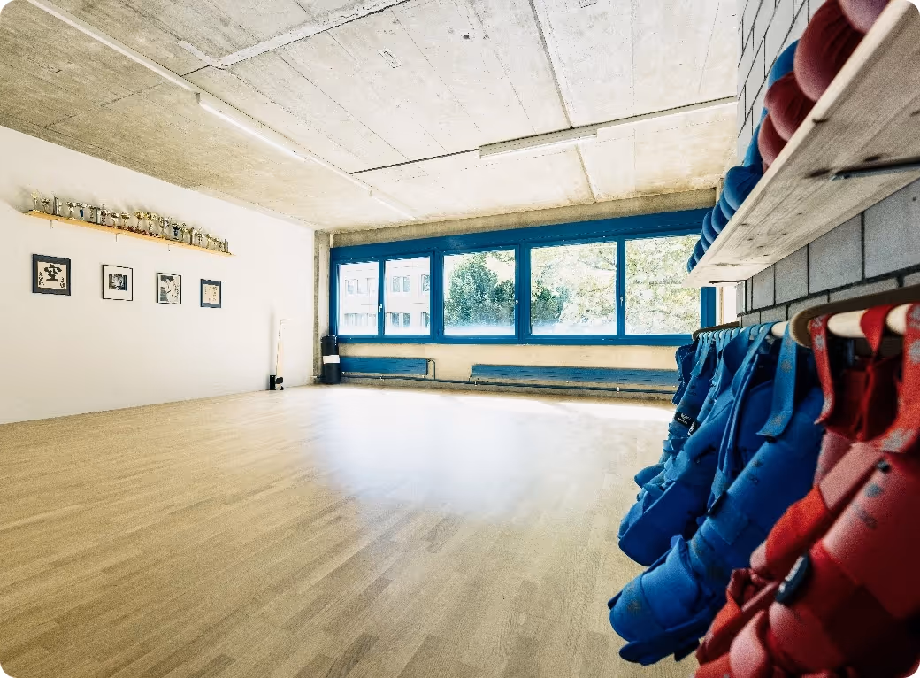 Bright martial arts dojo with wooden floor, blue window frames, and red and blue protective gear hanging on a wall rack.