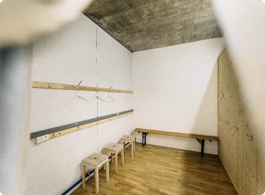 Minimalist changing room with wooden floor, three small wooden stools, a long wooden bench, white walls, and hangers on two wooden racks.