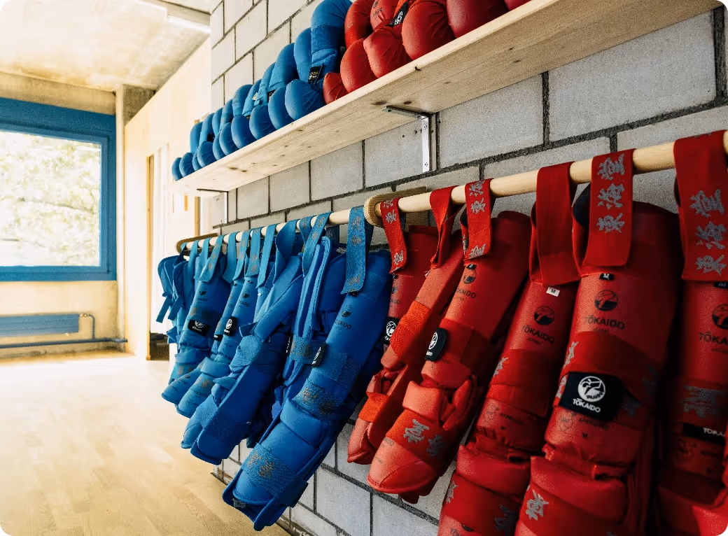Rows of red and blue protective martial arts gloves hanging on wooden rods in a bright training room with brick walls and large window.