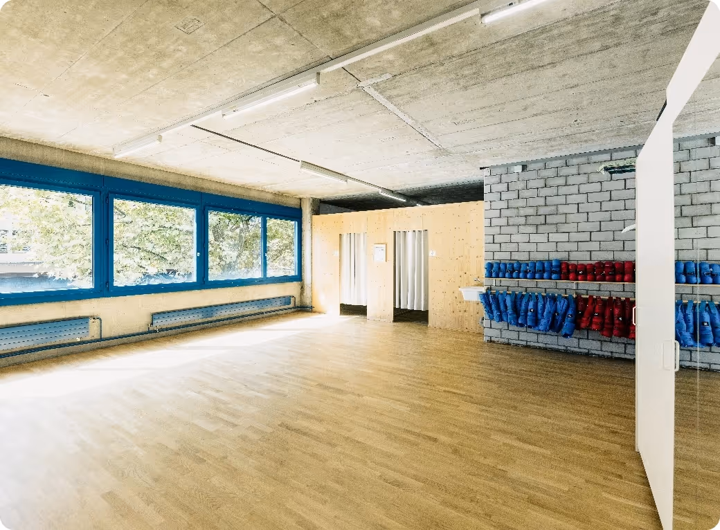 Bright room with large blue-framed windows, wooden floor, changing rooms with curtains, and shelves holding blue and red martial arts protective gear.