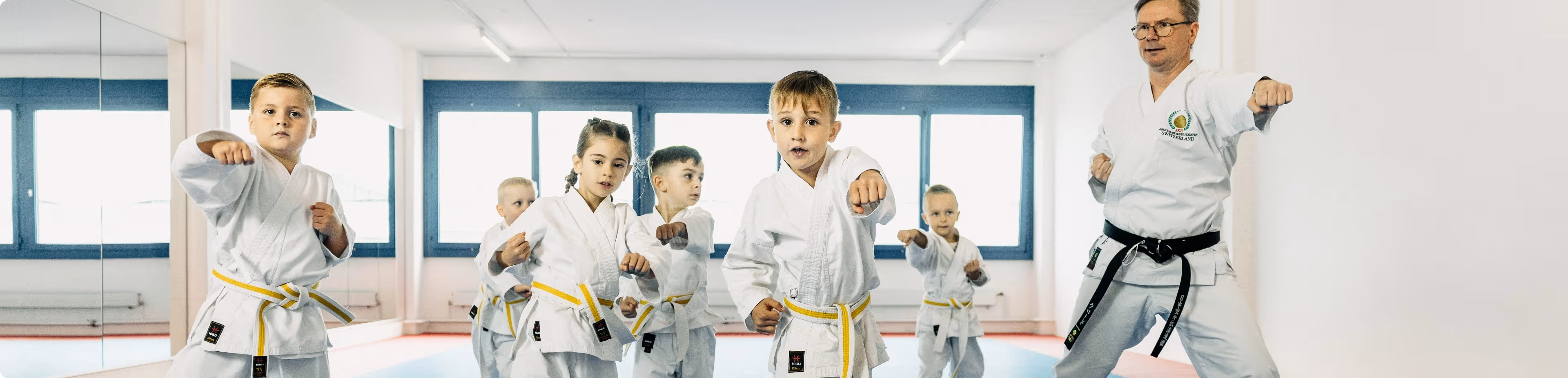 Group of children in white karate uniforms with yellow belts practicing punches with an instructor in a white uniform and black belt in a bright dojo.