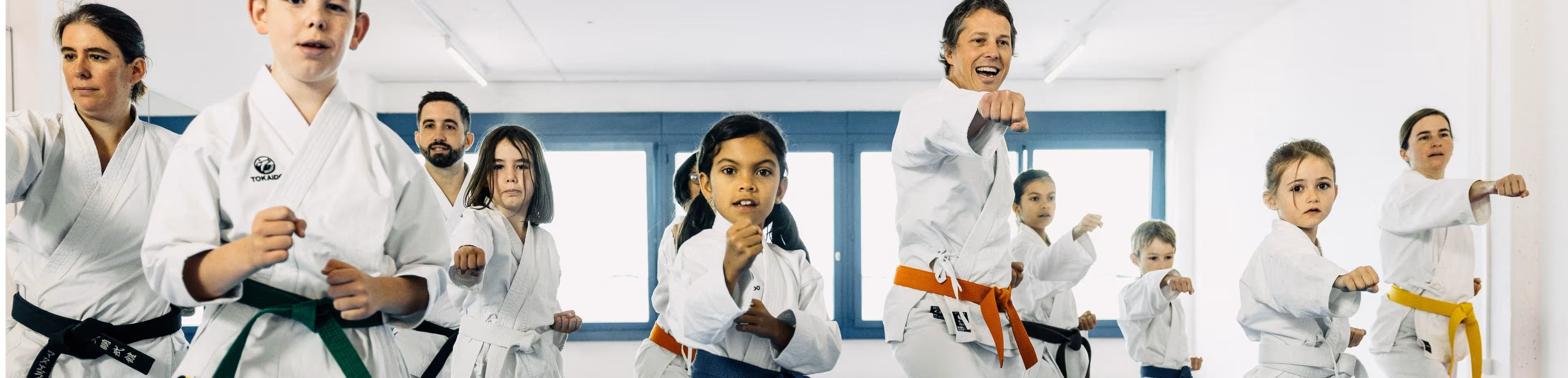 Group of children and adults in white karate uniforms performing punches in a dojo.