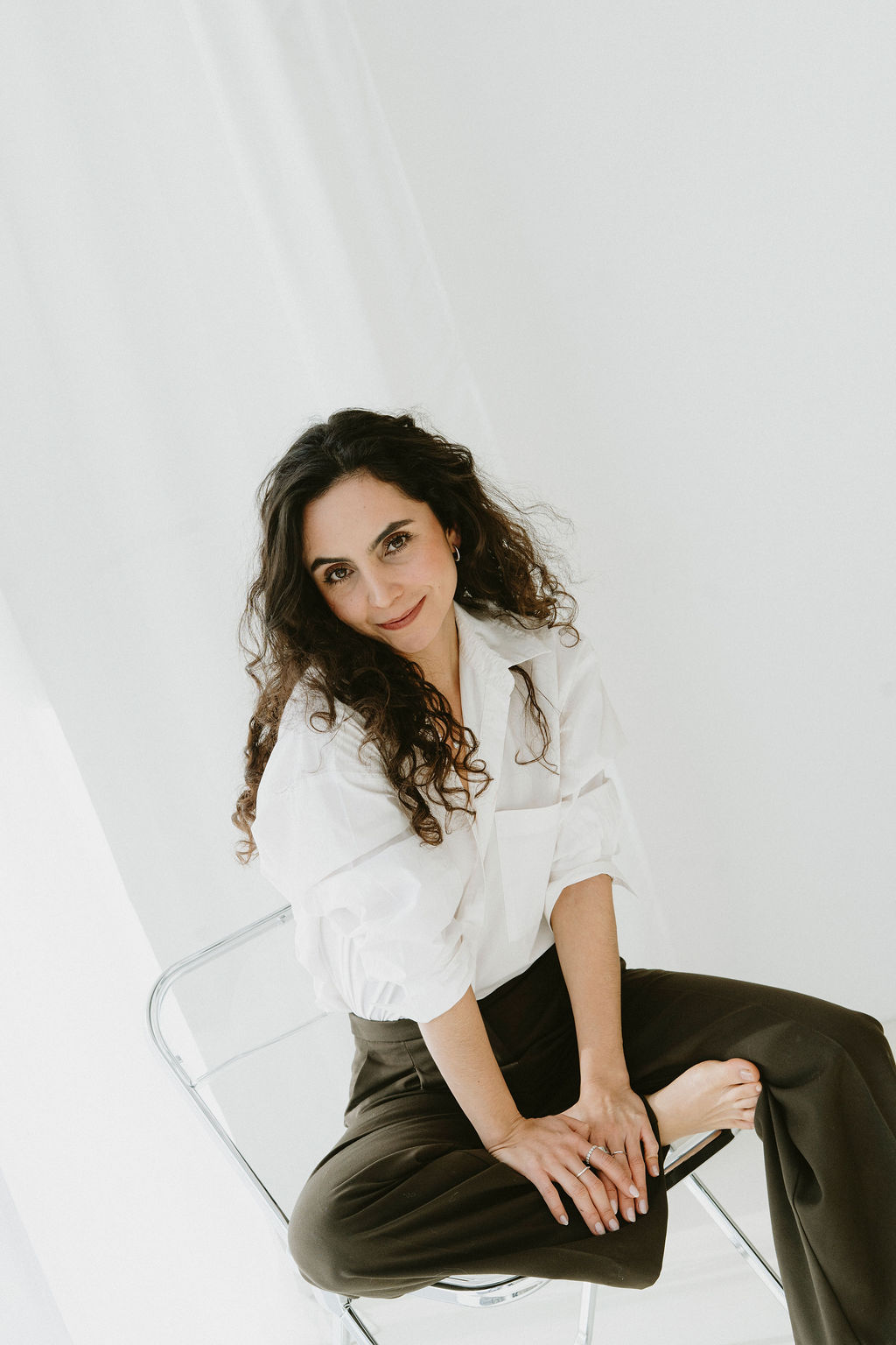 Smiling woman with curly hair sitting barefoot cross-legged on a transparent chair wearing a white shirt and dark pants.