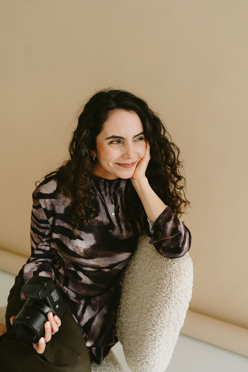 Smiling woman with curly hair holding a camera and leaning on a textured cream chair against a beige background.